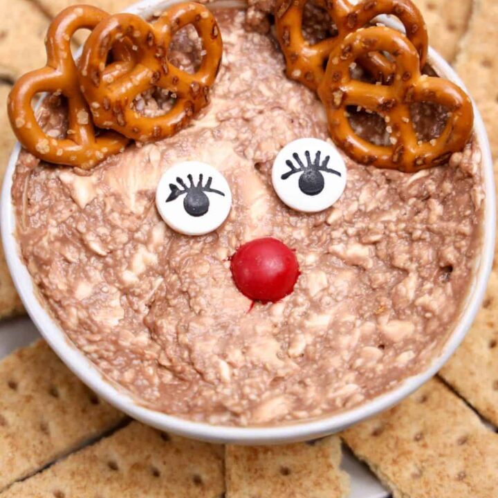 Rudolph the Reindeer Christmas Dip in a white bowl, decorated with candy eyes, a red M&M nose, and mini pretzels as antlers. The Nutella and cream cheese mixture is surrounded by neatly arranged graham cracker pieces for dipping.