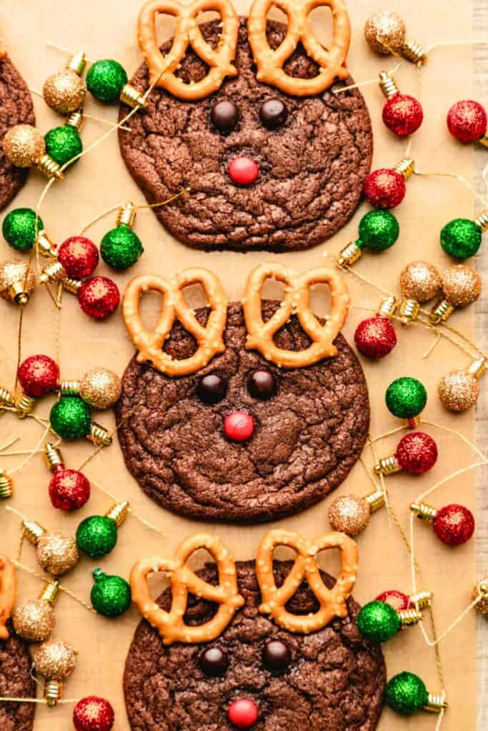 Chocolate Christmas cookies decorated to look like reindeer, with pretzel twists for antlers, chocolate candies for eyes, and a red candy nose. The cookies are arranged on parchment paper surrounded by festive mini ornaments in red, green, and gold.