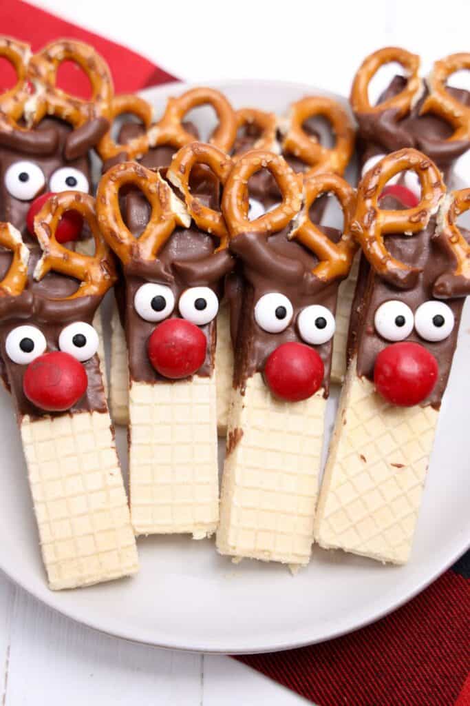 Close-up of reindeer cookies made from vanilla wafer bars dipped in chocolate, topped with candy eyes, red candy noses, and mini pretzels arranged as antlers. The cookies are neatly lined on a white plate with a red and black cloth underneath, emphasizing their playful holiday design.