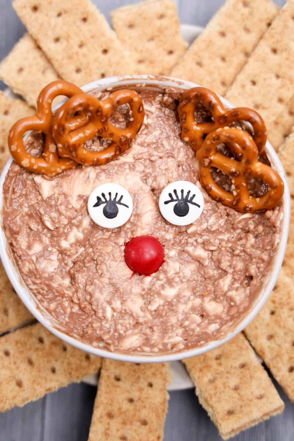 Reindeer cream cheese dip decorated with candy eyes and a red candy nose with mini pretzels arranged as antlers. Graham cracker sticks surround the bowl for dipping a sweet Christmas Eve snack.
