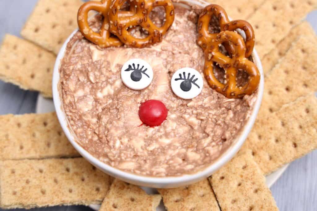Holiday dip styled as a reindeer with pretzel antlers, candy eyes with lashes, and a red M&M nose, served in a white bowl. The creamy chocolate dip is surrounded by graham cracker pieces, making it a festive and fun addition to holiday dips and appetizers.
