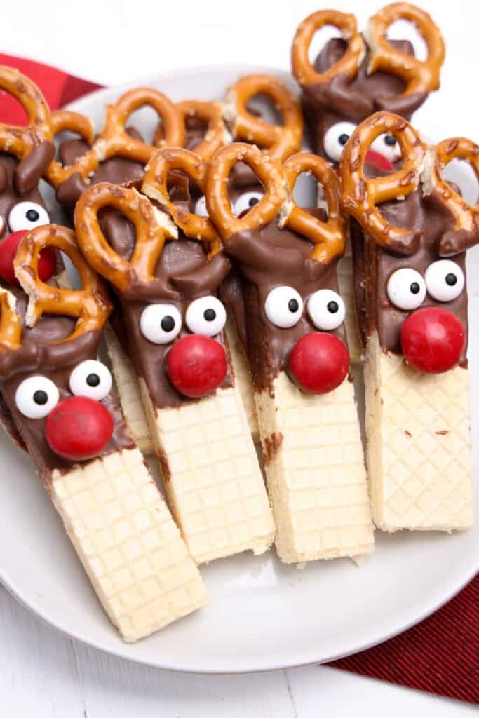 Close up of reindeer cookies made from vanilla wafer bars dipped in chocolate, topped with candy eyes, red candy noses, and mini pretzels arranged as antlers. The cookies are neatly lined on a white plate with a red and black cloth underneath.