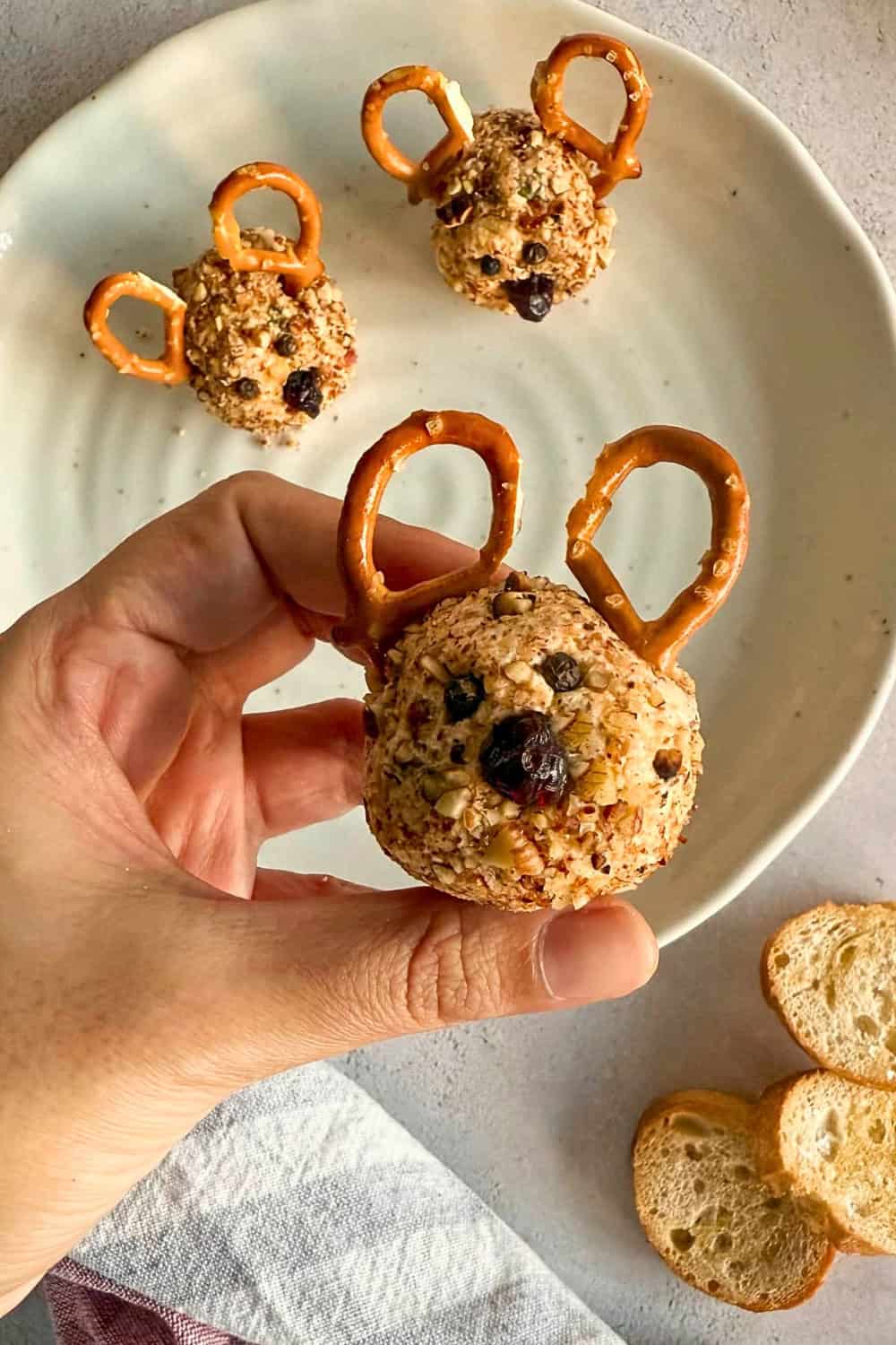 Reindeer mini cheese balls coated with chopped nuts and decorated with pretzel antlers. Small dark pieces form the eyes and nose with one cheese ball held in a hand above a plate.