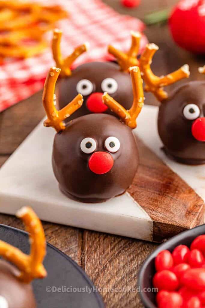Chocolate-coated Oreo balls are decorated as reindeer with candy eyes, red M&M noses, and pretzel antlers. These festive reindeer Oreo balls are displayed on a marble and wood serving board, ready to be used as a fun holiday dessert.