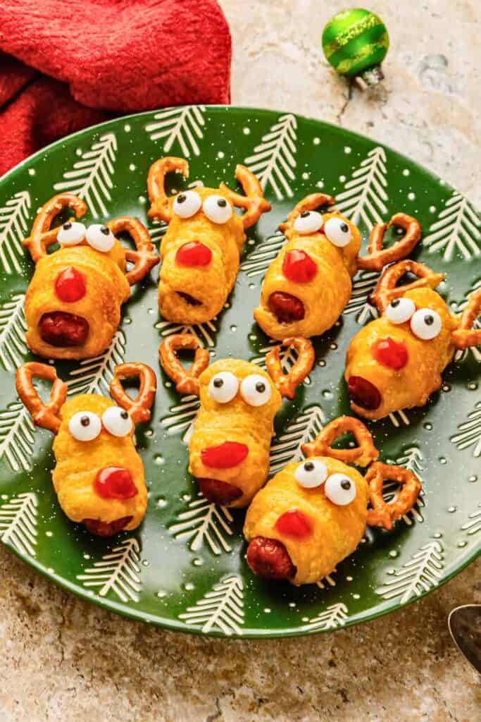 Reindeer pigs in a blanket made with sausages wrapped in golden pastry and decorated with pretzel antlers, candy eyes, and red noses. The festive Christmas appetizers are arranged on a green holiday plate.