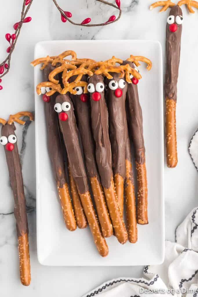 Chocolate-dipped pretzel rods are decorated to look like reindeer using candy eyes, red M&M noses, and broken pretzel pieces as antlers. These festive reindeer pretzel rods are arranged on a white rectangular plate, perfect for an easy holiday treat.