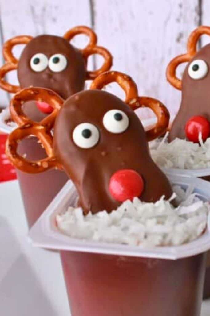 Chocolate pudding cups are topped with shredded coconut and decorated with reindeer made from chocolate-covered cookies, candy eyes, red M&M noses, and pretzel antlers, a fun holiday snack for kids.