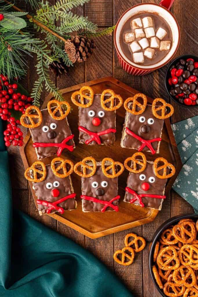 Festive reindeer Rice Krispie treats coated in chocolate and decorated with candy eyes, red or brown noses, red licorice scarves, and pretzel antlers. Arranged on a wooden tray with hot cocoa, pine sprigs, and bowls of candy for a playful holiday snack display.