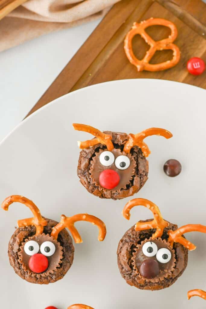 Close-up of three reindeer snack brownies on a white plate, each decorated with pretzel antlers, candy eyes, Reese’s mini cups, and red or brown M&M noses. A whole pretzel and a red M&M are seen on a wooden board in the background.
