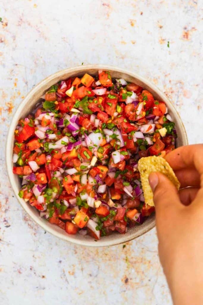 Fresh Restaurant-Style Pico De Gallo in a white bowl, made with diced tomatoes, red onions, jalapeños, cilantro, and garlic. A hand dips a tortilla chip into the colorful salsa, highlighting its chunky, vibrant texture.