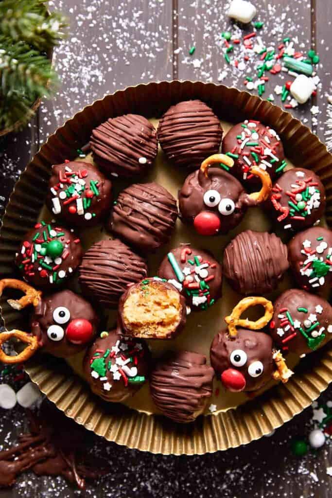 Festive no bake Rice Krispie peanut butter balls coated in chocolate, some topped with holiday sprinkles and others decorated as reindeer with candy eyes, red noses, and pretzel antlers. One ball is cut open to show the crispy peanut butter center.
