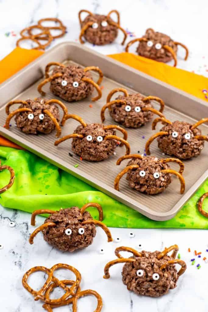 Chocolate Rice Krispie spider treats with candy eyes and pretzel stick legs arranged on a baking sheet and table, with colorful napkins and scattered sprinkles.