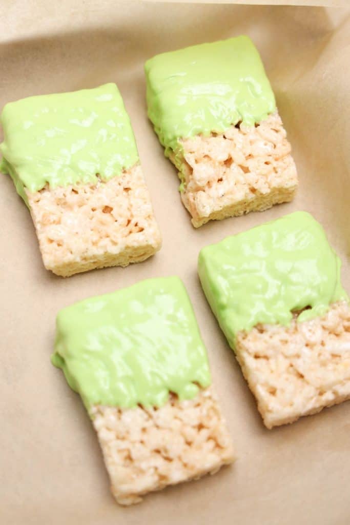 Four Rice Krispie treats placed on parchment paper, each partially dipped in smooth green candy coating. These undecorated treats are in the early stages of being transformed into Halloween-themed snacks.