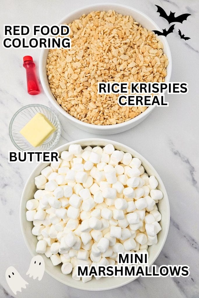 Overhead view of Halloween-themed rice krispie treat ingredients including a bowl of rice krispies cereal, a bowl of mini marshmallows, a pat of butter in a small glass dish, and a bottle of red food coloring, decorated with ghost and bat graphics.