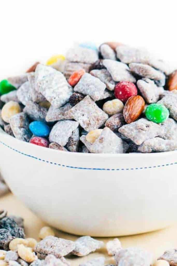 A white bowl filled with trail mix muddy buddies coated in powdered sugar mixed with colorful candy coated chocolates almonds and crunchy cereal pieces. Some of the snack mix spills onto the surface around the bowl highlighting texture and variety in this sweet and crunchy road trip treat.