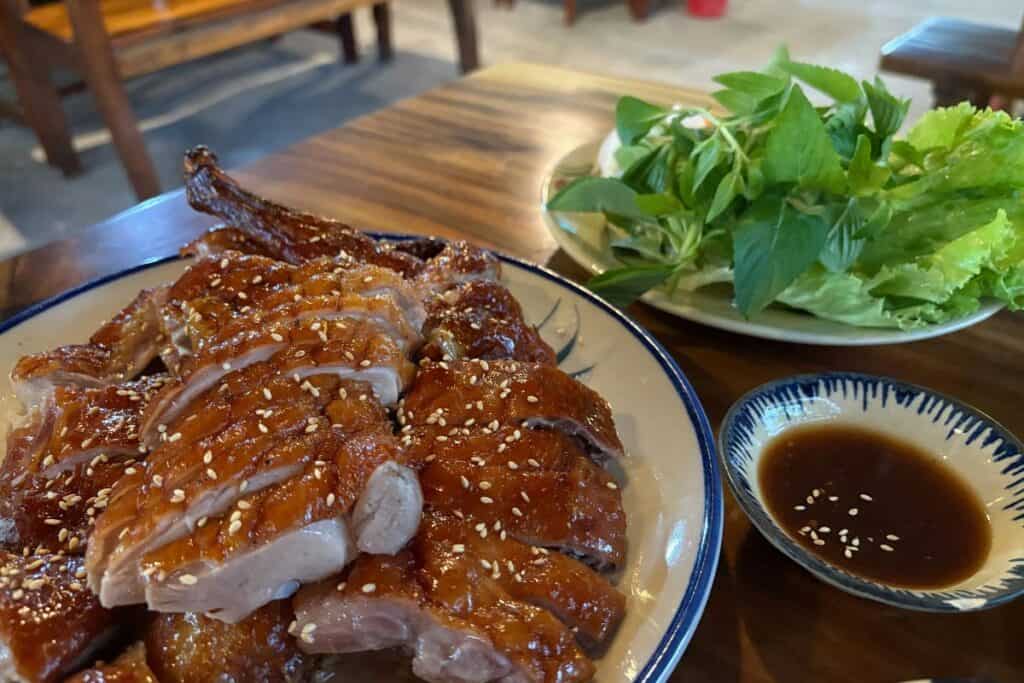 Close up of roast duck cut into pieces on a plate with dipping sauce and green veggies in th background. 