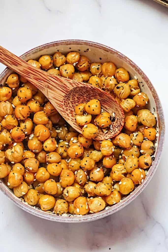 A bowl of roasted seasoned chickpeas coated in oil and spices with visible black and white seeds mixed throughout and a wooden spoon resting inside.