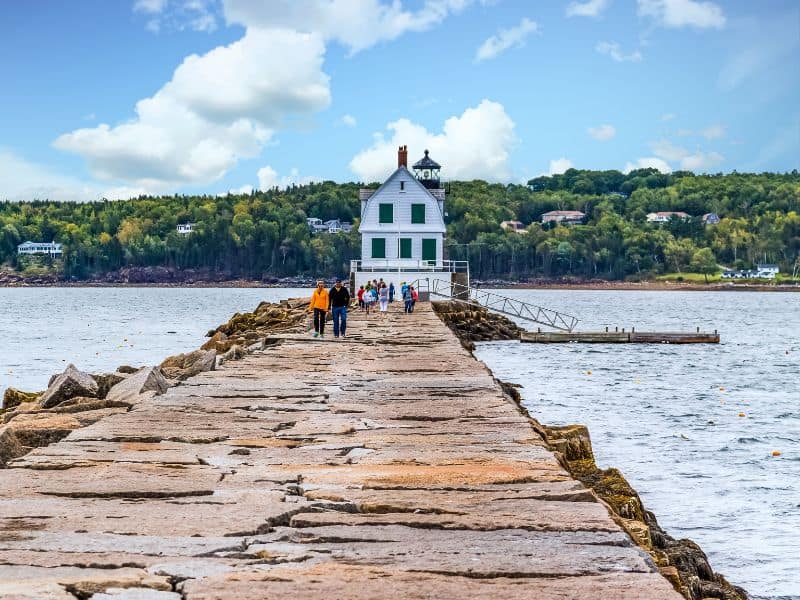 People walking along a stone breakwater to the old Rockland Breakwater Lighthouse in Rockland, ME. Trees and clouds across the water. 
