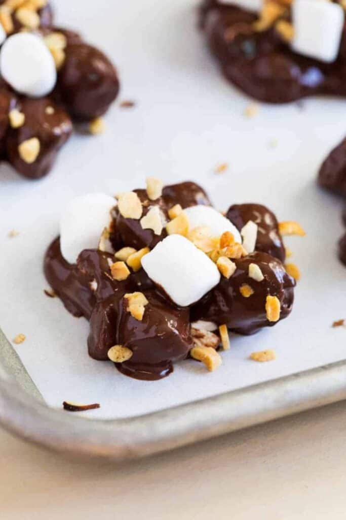 Clusters of rocky road candy coated in glossy chocolate and topped with mini marshmallows and crushed nuts on a parchment lined baking tray. The close up view highlights the chunky texture and combination of chocolate, marshmallows, and crunchy mix ins in this no bake sweet treat.