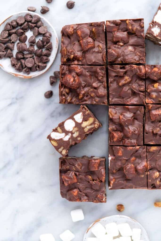 Rocky road fudge cut into squares, packed with mini marshmallows, chopped nuts, and chocolate chunks. Chocolate chips and marshmallows are scattered nearby on a marble surface for a cozy, homemade feeling.