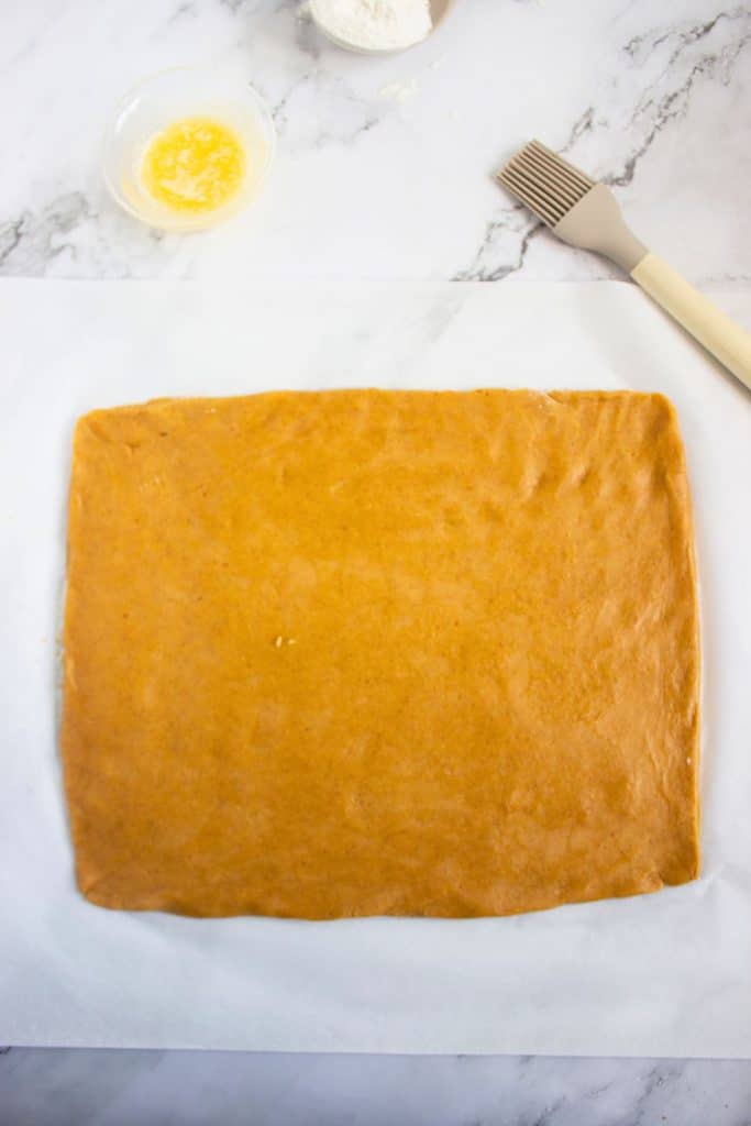 Pumpkin cookie dough rolled into a large, even rectangle on parchment paper, ready to be filled or shaped. A bowl of melted butter, flour, and a pastry brush sit nearby on a marble countertop.
