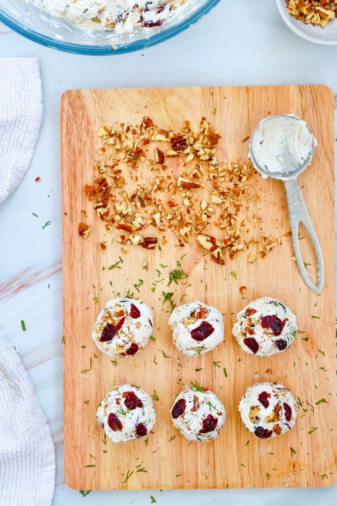Overhead view of six round cranberry cheese balls arranged on a wooden cutting board with dried cranberries, chopped pecans, and fresh dill visible throughout. A metal measuring cup filled with the cheese mixture rests beside a pile of chopped pecans, showing how the mixture is portioned before shaping.