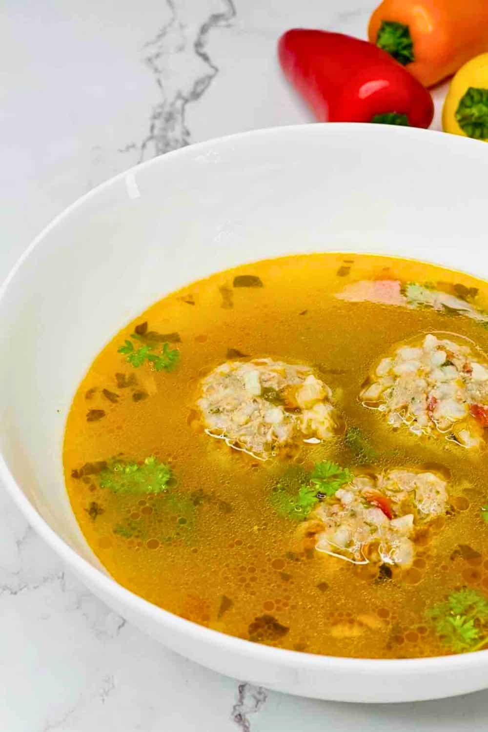 Romanian meatball soup with tender meatballs and fresh parsley is served in a clear golden broth. Colorful bell peppers in the background hint at the vegetables used in the soup.