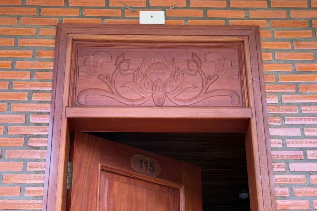 Wooden doorway of Room 115 at Palm Bay Resort in Phu Quoc, Vietnam, featuring an intricate carved lotus flower design above the door set against a brick wall background.