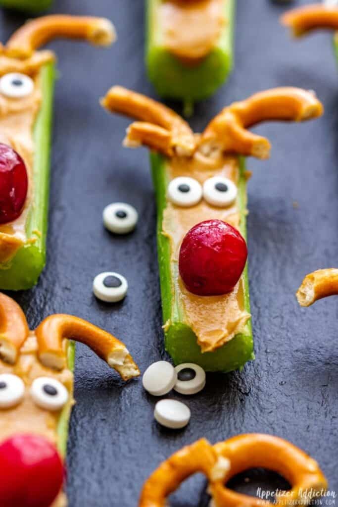 Celery sticks topped with peanut butter are decorated to look like reindeer, using candy eyes, a red grape or cranberry for the nose, and broken pretzels for antlers. These festive reindeer snacks for kids are arranged on a dark slate surface, with extra candy eyes scattered around.
