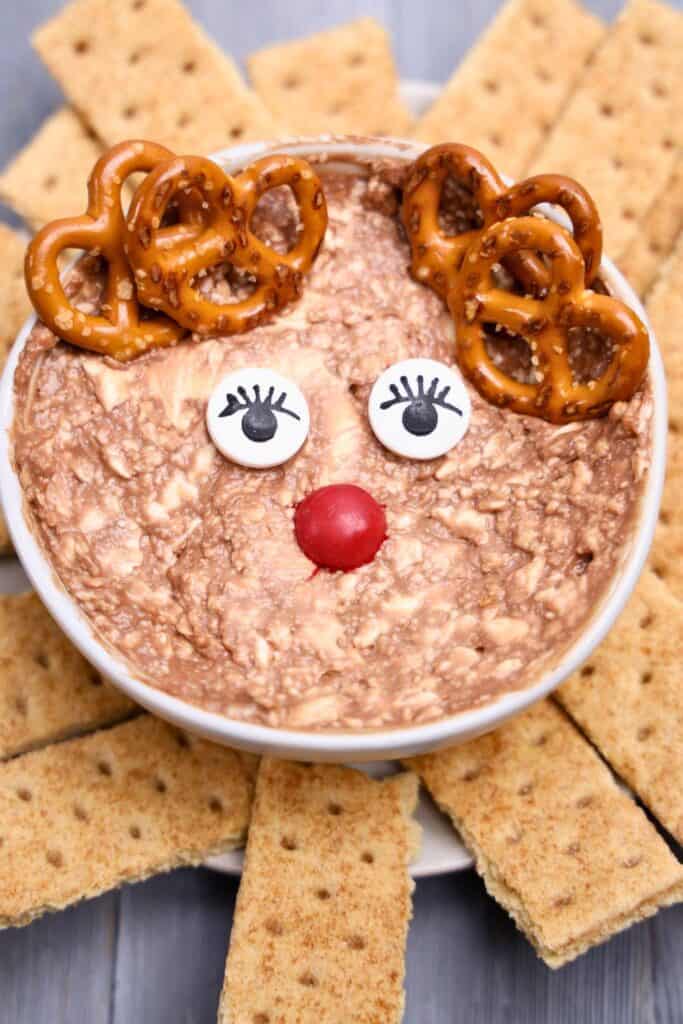Rudolph Christmas Dip presented in a white bowl, decorated to resemble a reindeer with candy eyes, a red M&M nose, and pretzel antlers. The creamy chocolate dip is surrounded by neatly arranged graham cracker pieces for dipping.