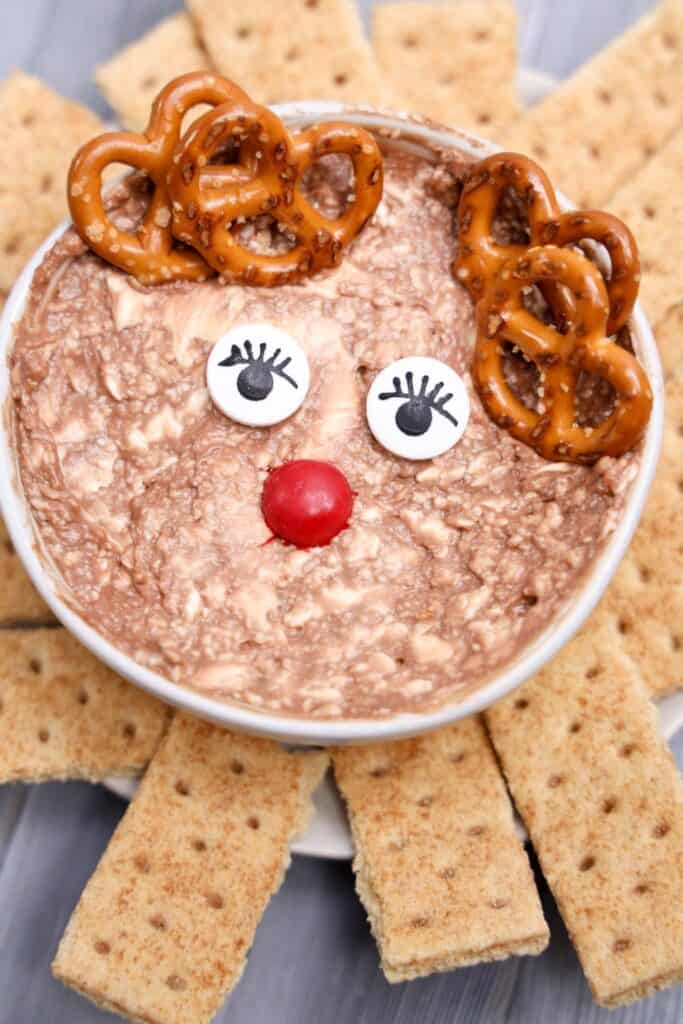 Festive Rudolph snack dip served in a white bowl, decorated to look like a reindeer with pretzel antlers, candy eyes with lashes, and a red M&M nose. The creamy chocolate dip is surrounded by rectangular graham crackers for dipping.