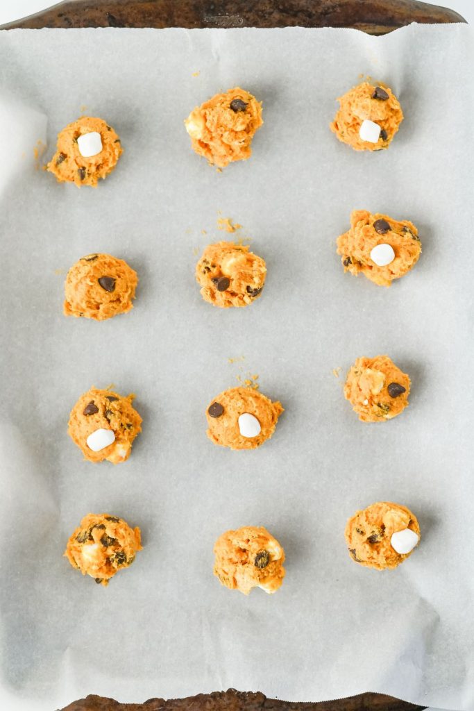 Overhead view of unbaked s’mores cookie dough scoops on a parchment-lined baking sheet. The orange dough is studded with chocolate chips and mini marshmallows, ready to bake as part of a cozy s’mores dessert recipe.