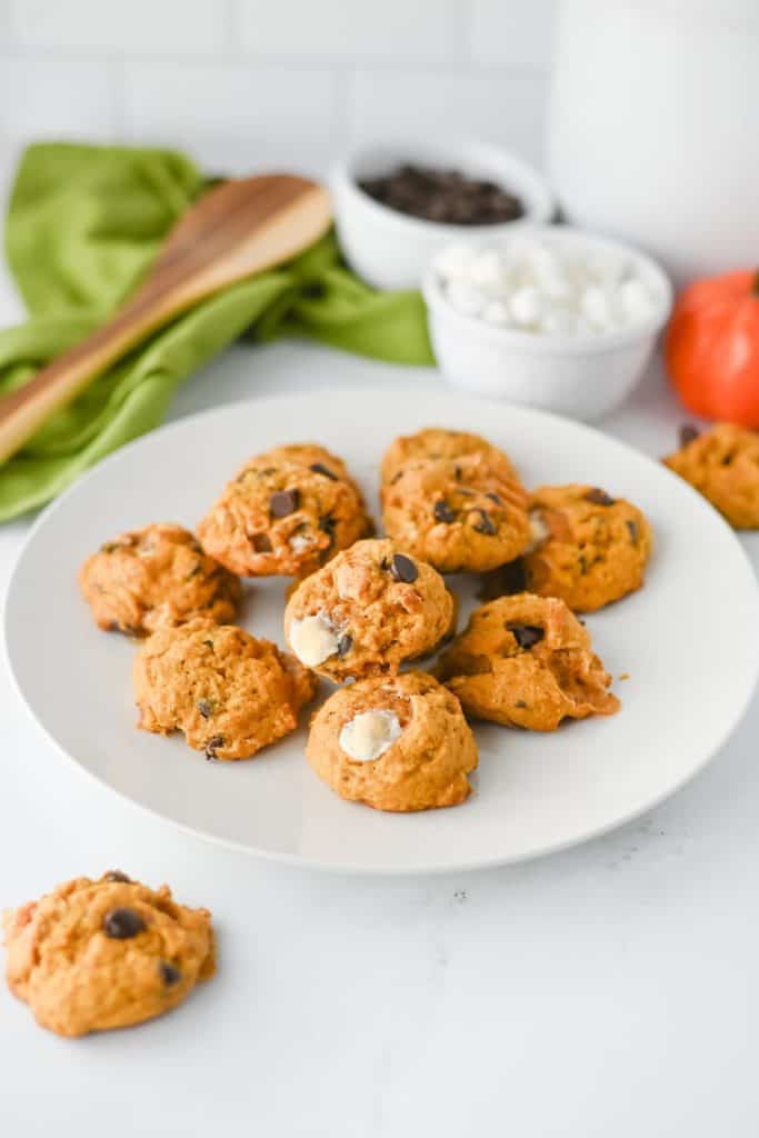 Plate of freshly baked s’mores cookies with visible chocolate chips and toasted marshmallow centers. A green cloth, wooden spoon, and bowls of mini marshmallows and chocolate chips are styled in the background.