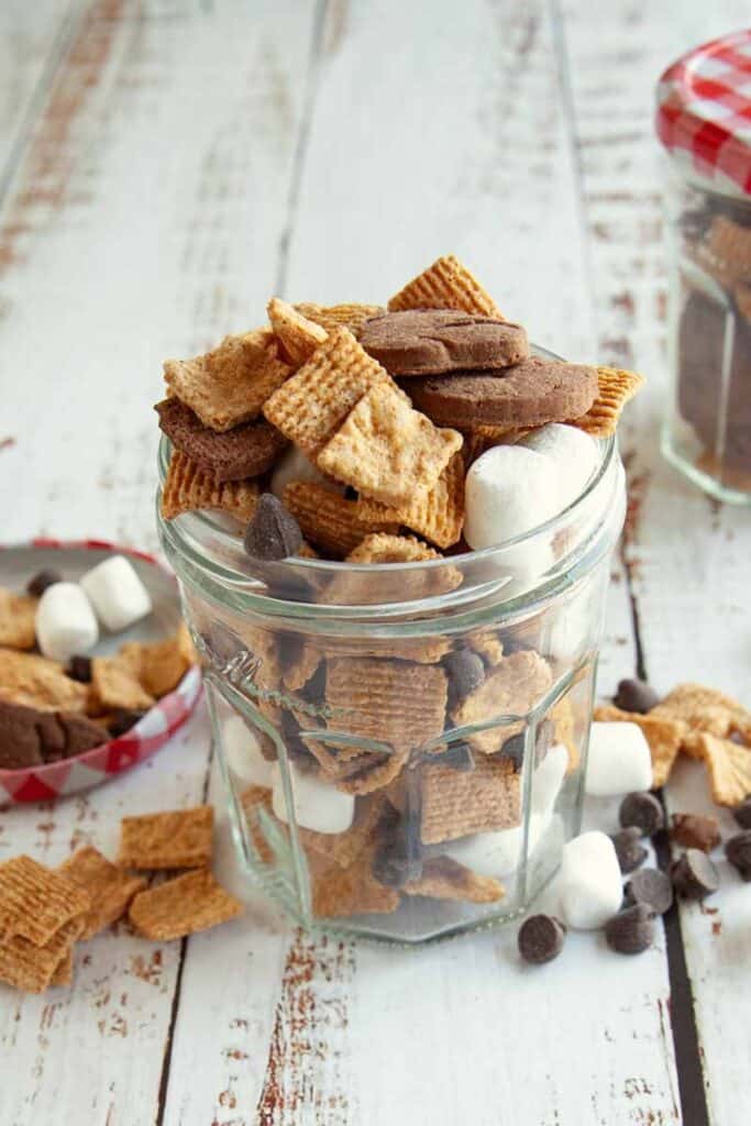 Glass jar filled with a s&rsquo;mores snack mix made of graham cereal squares, mini marshmallows, chocolate chips, and chocolate cookies. Extra marshmallows and chocolate chips are scattered on the white wooden table around the jar.