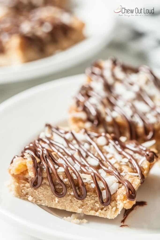 A slice of samoas cookie bars recipe served on a plate with a crumbly shortbread base topped with caramel coconut and drizzled with chocolate. Another slice sits blurred in the background showing the layered texture of this rich dessert.