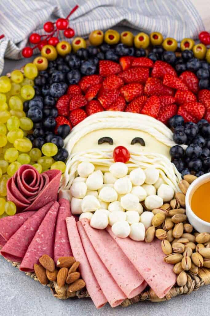 A Santa-themed charcuterie board styled with mozzarella balls for the beard, string cheese and olives for facial features, and a cherry tomato nose, surrounded by strawberries, blueberries, grapes, olives, deli meats, nuts, and a cup of honey for dipping, making it a fun and savory twist on Christmas movie night snacks.