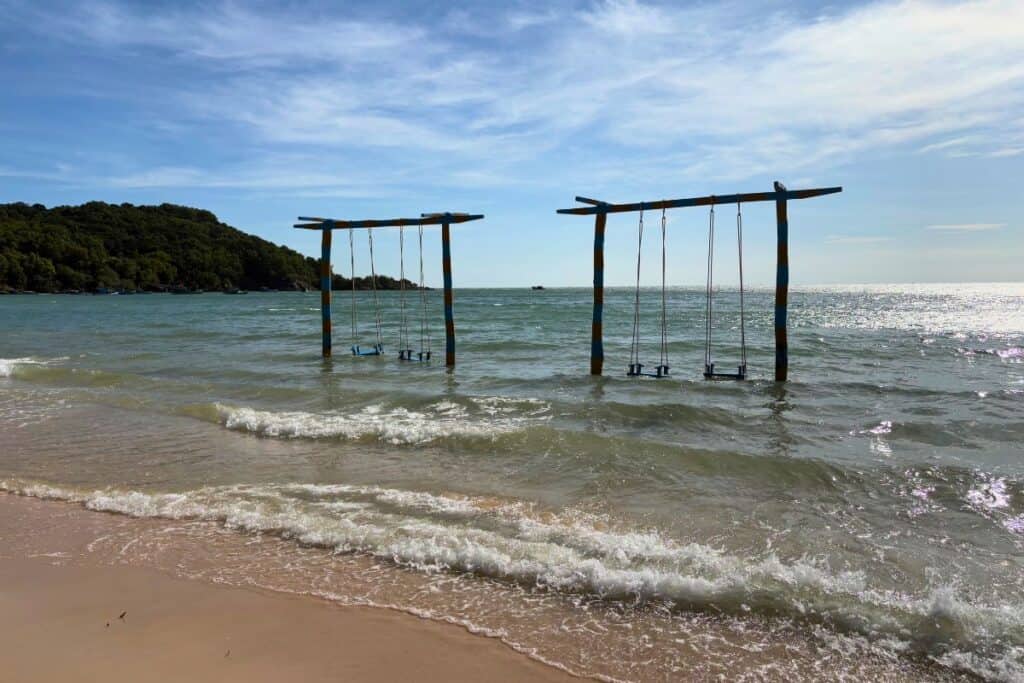 Two sets of wooden swings standing in shallow ocean water at Sao Beach, Phu Quoc, Vietnam, with gentle waves lapping at the sandy shore and a forested hill in the background under a blue sky