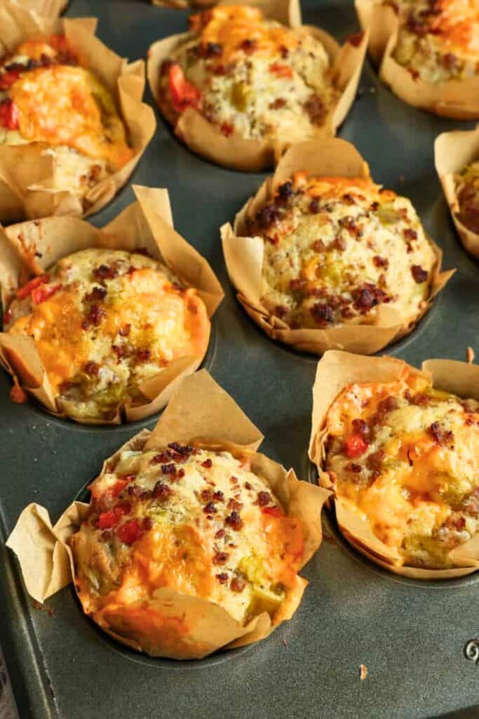 Tray of golden baked muffins in parchment liners filled with melted cheese sausage and bits of peppers. These on the go breakfast muffin with sausage and cheese are shown fresh from the oven with crispy tops and a soft savory interior.