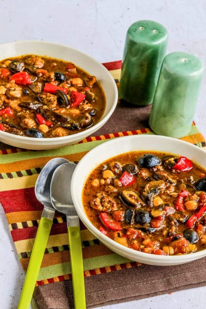 Two white bowls of hearty Sausage and Chickpea Stew filled with browned sausage, chickpeas, red peppers, mushrooms, black olives, and tomatoes in a rich, savory broth.