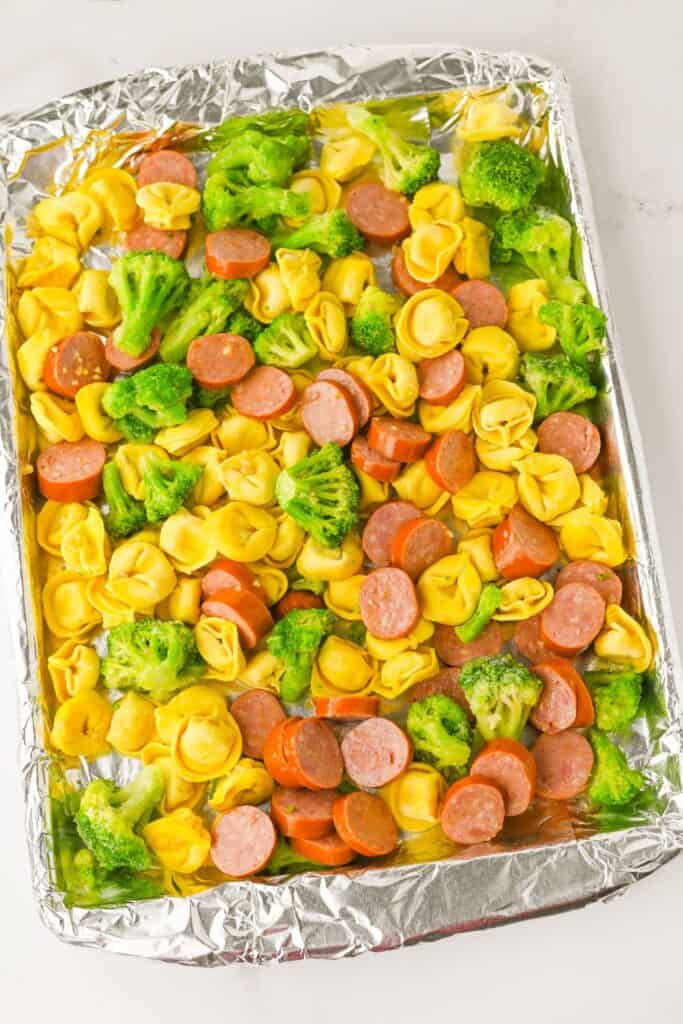 Foil-lined sheet pan filled with uncooked tortellini, sliced sausage, and broccoli florets, evenly spread and ready for the oven.