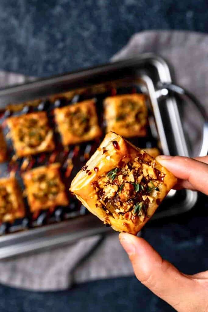 A hand holds a golden savory pear tartlet topped with chopped nuts and fresh herbs. More square puff pastry tartlets are arranged on a baking tray in the background