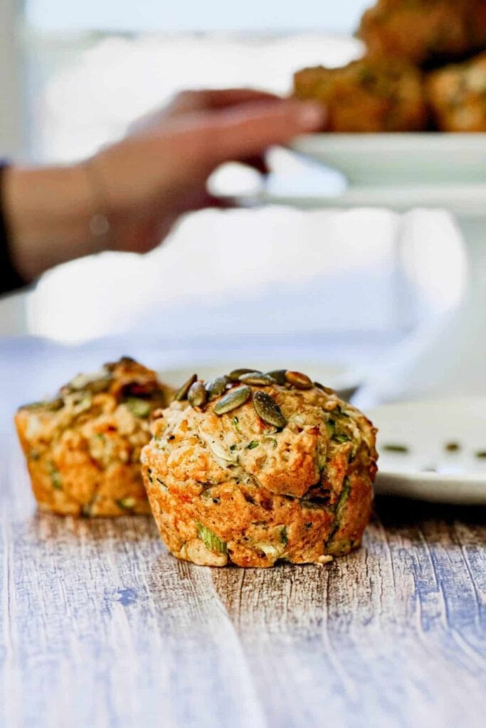 Savory vegan zucchini muffins with a golden textured top sprinkled with seeds and visible green zucchini pieces baked throughout. Blurred in the background is a hand reaching for one. 
