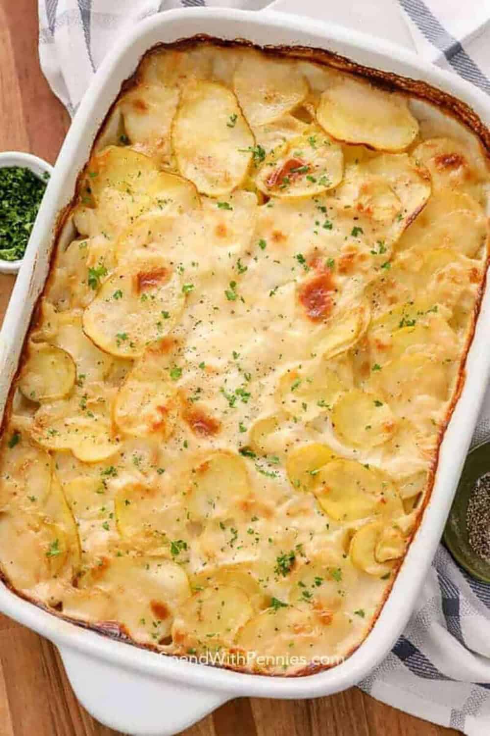 A white baking dish filled with creamy scalloped potatoes, layered with thinly sliced potatoes in a rich sauce and baked until golden brown and bubbling around the edges, finished with chopped fresh parsley and black pepper.
