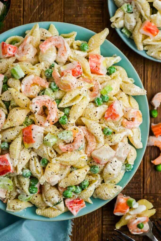 Creamy seafood pasta salad served on a teal plate, featuring shell pasta mixed with shrimp, imitation crab, green peas, celery, and diced red bell pepper. The salad is coated in a rich, pepper-speckled dressing.