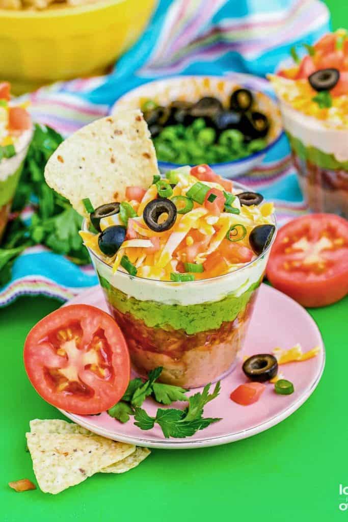 Colorful Seven Layer Taco Dip served in a clear cup, showing layers of refried beans, salsa, guacamole, sour cream, shredded cheese, diced tomatoes, and black olives. Garnished with green onions and a tortilla chip for dipping, with fresh tomatoes on the side.
