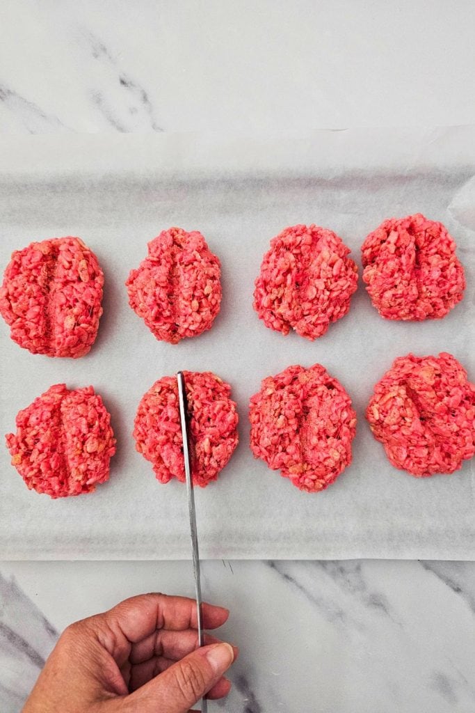 Hand using a knife to press a center groove into bright red rice krispie treat balls on a parchment-lined baking sheet, shaping them to look like brains for Halloween.
