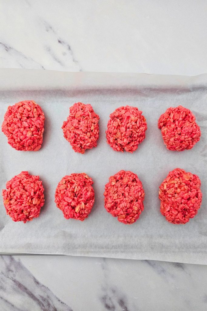 Eight scoops of bright red rice krispie treats arranged on a parchment-lined baking sheet, cooling before being shaped into Halloween-themed brain designs.