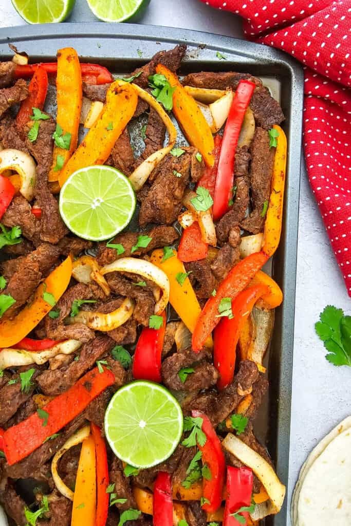 Sheet pan beef fajitas feature seasoned strips of steak roasted with red and orange bell peppers and sliced onions. Fresh cilantro and lime halves top the fajitas with flour tortillas served alongside the pan.