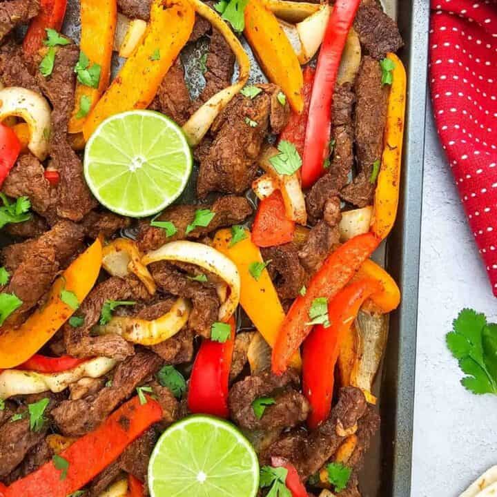 Sheet pan beef fajitas feature seasoned strips of steak roasted with red and orange bell peppers and sliced onions. Fresh cilantro and lime halves top the fajitas with flour tortillas served alongside the pan.