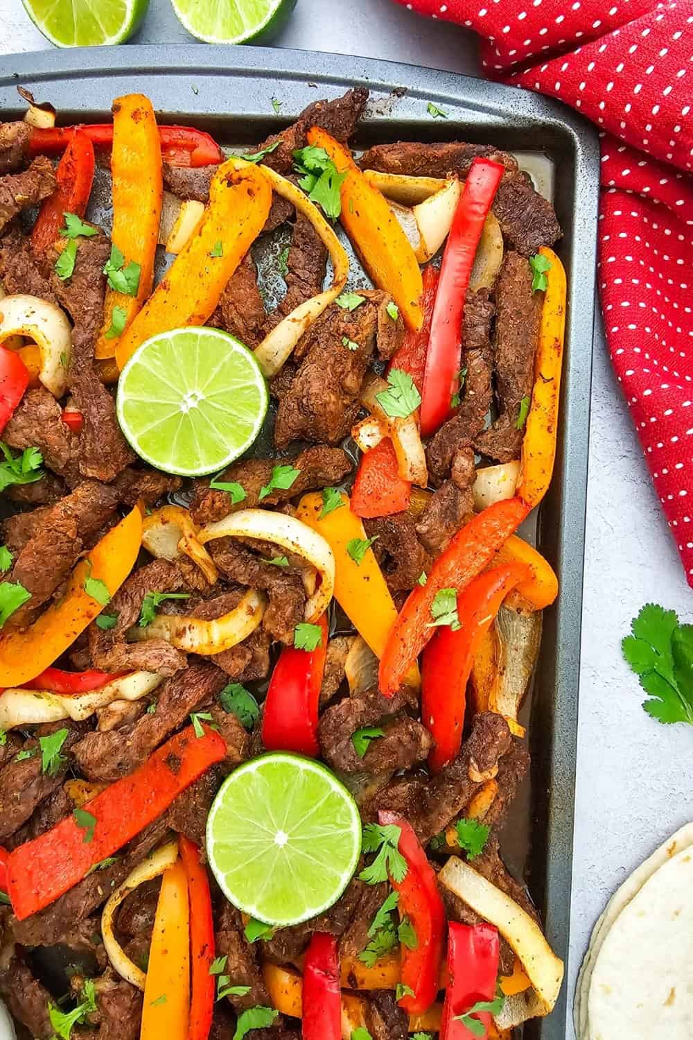 Sheet pan beef fajitas feature seasoned strips of steak roasted with red and orange bell peppers and sliced onions. Fresh cilantro and lime halves top the fajitas with flour tortillas served alongside the pan.